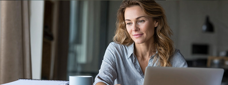 A focused professional studying for an online MBA in a modern workspace, engaging with course materials on a laptop surrounded by study notes.